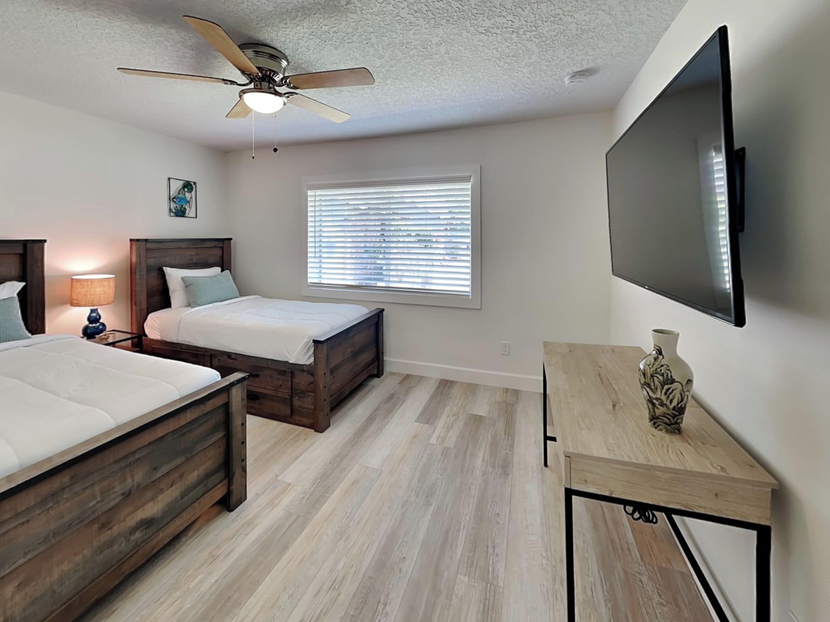 A bedroom with two beds, a ceiling fan, a TV, a window with blinds, and a small table with a vase.