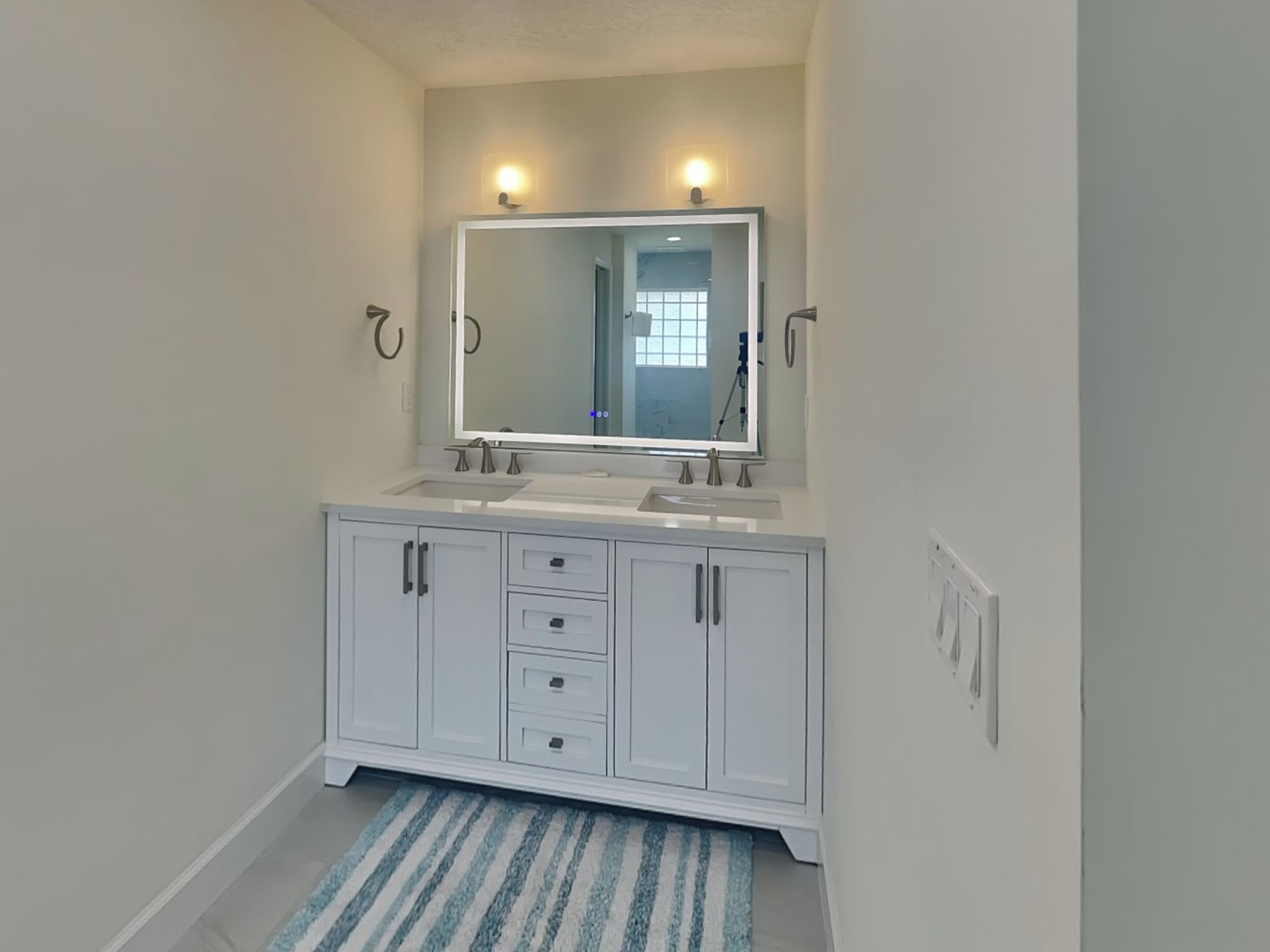 A bathroom with a double sink vanity, a large mirror, and a striped rug on the floor.