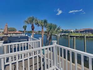 A waterfront deck with white railings, palm trees, and a view of the water with boats and buildings in the background. - Dolphin View Oasis by Distinctive Beach Rentals (Palm Coast)