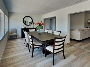 Une salle à manger avec une longue table, six chaises, un vase de fleurs, un miroir et une porte donnant sur une autre pièce.