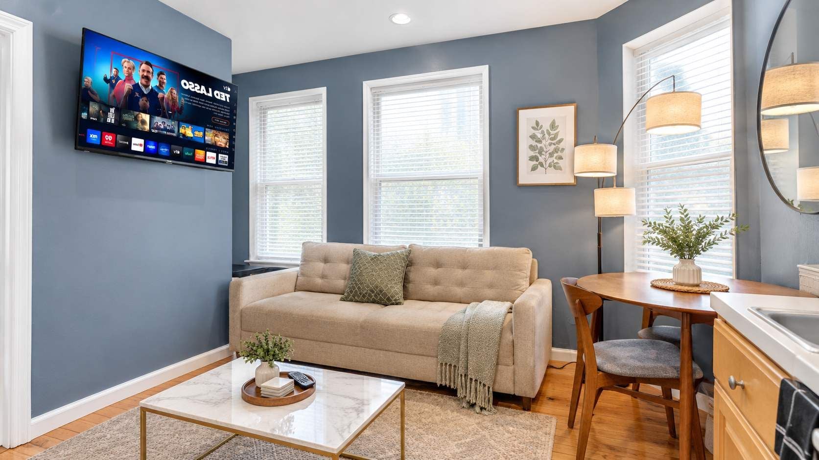A living room with a beige sofa, a coffee table, a TV mounted on the wall, and a window with blinds.