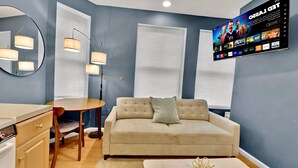 A modern living room with a beige sofa, a round dining table, and a TV mounted on the wall displaying a movie. - Historic Rittenhouse 2BR Loft | Sleeps 12 | Philly (Philadelphia)