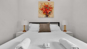 A hotel room with a bed, white linens, a brown pillow, two bedside lamps, and a painting of flowers on the wall.