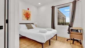 Une chambre d’hôtel avec un lit, un bureau, une fenêtre avec des rideaux, un tableau au mur et un plancher en bois.