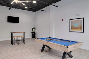 A pool table in a room with a wooden floor, a small table with stools, a mounted TV, and a framed picture on the wall.