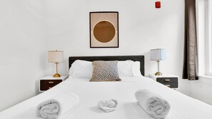 A hotel room with a neatly made bed, two bedside tables with lamps, a framed artwork above the bed, and a window with curtains.