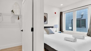 A hotel room with a bed, a window with curtains, a bedside table with a lamp, and a wall-mounted shelf with a decorative item.