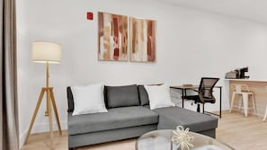 A modern living room with a grey sofa, a floor lamp, a coffee machine, and a desk with two chairs.