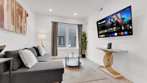 A modern living room with a grey sofa, a glass coffee table, a flat-screen TV mounted on the wall, and a potted plant.