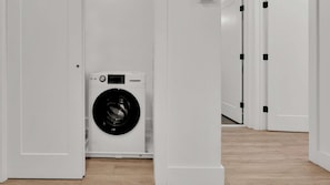 A laundry room with a front-loading washing machine, white walls, and a wooden floor.