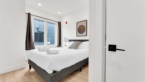 A modern hotel room with a large bed, a bedside table with a lamp, a window with curtains, and a framed picture on the wall.
