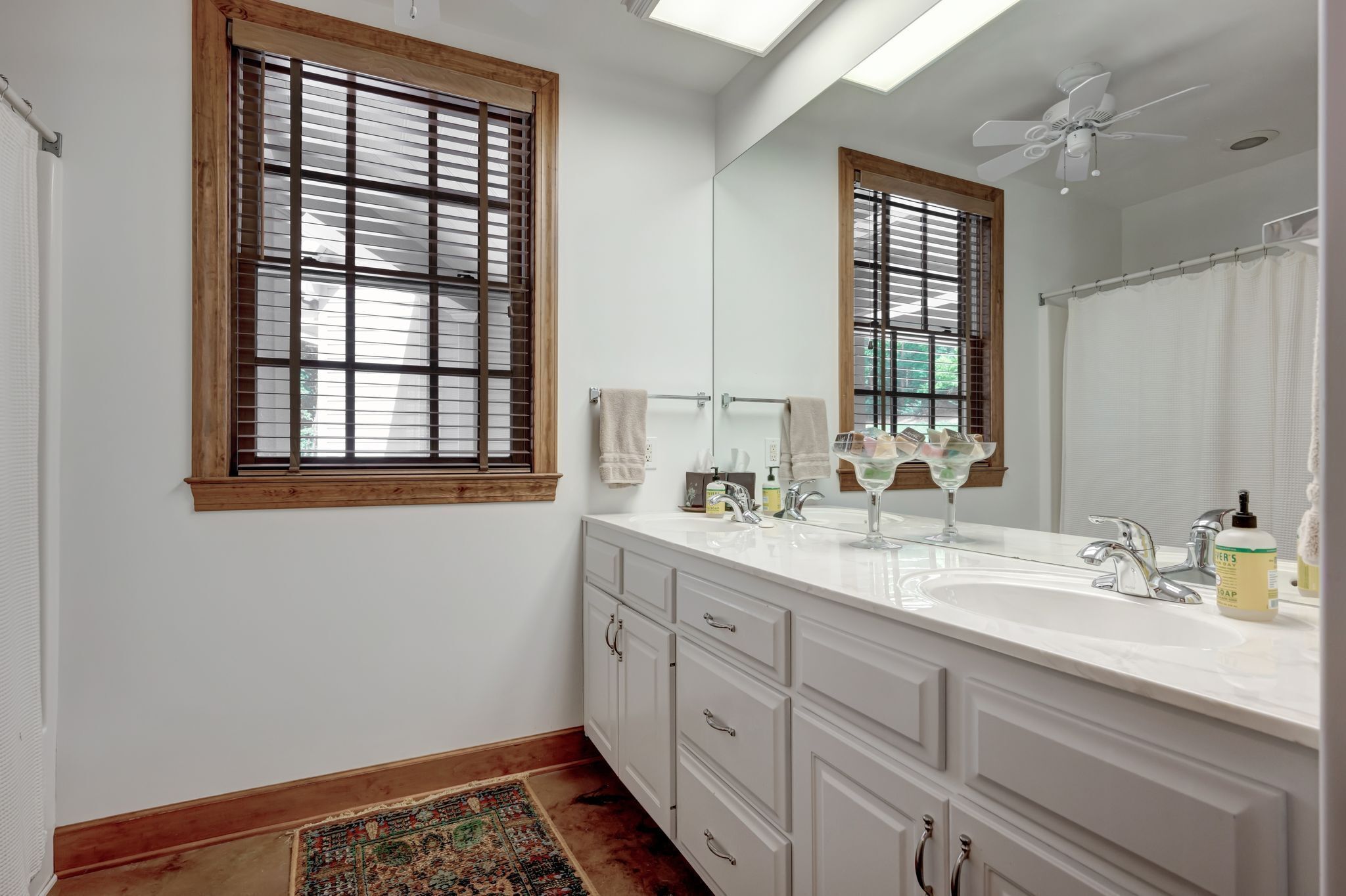 A bathroom with a large mirror, two sinks, a ceiling fan, and a window with blinds.