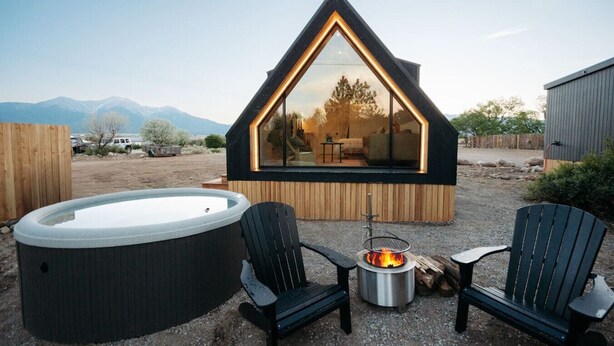 Un moderno hidromasaje triangular con dos sillas Adirondack y una chimenea en un entorno rústico al aire libre.