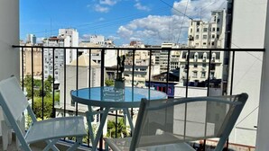 A balcony with a glass table and two chairs, overlooking a cityscape with various buildings.