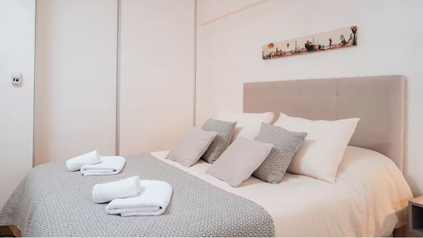 A neatly made bed with a grey patterned bedspread, multiple pillows, and folded towels on the floor.