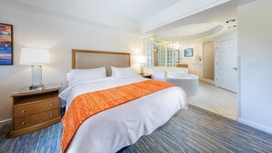 Ein Hotelzimmer mit einem großen Bett, einem Nachttisch, einer Lampe, einem Badezimmer mit einer Badewanne und einer Tür zu einem weiteren Raum.