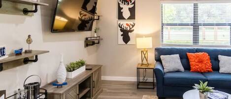 Un salón con un sofá azul, una mesa de centro, un televisor montado en la pared y una obra de arte que representa ciervos.