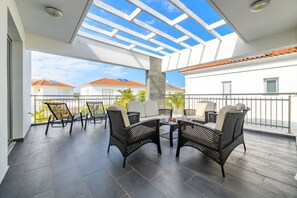 Een balkon met rieten meubilair, een glazen dak en uitzicht op omliggende huizen.