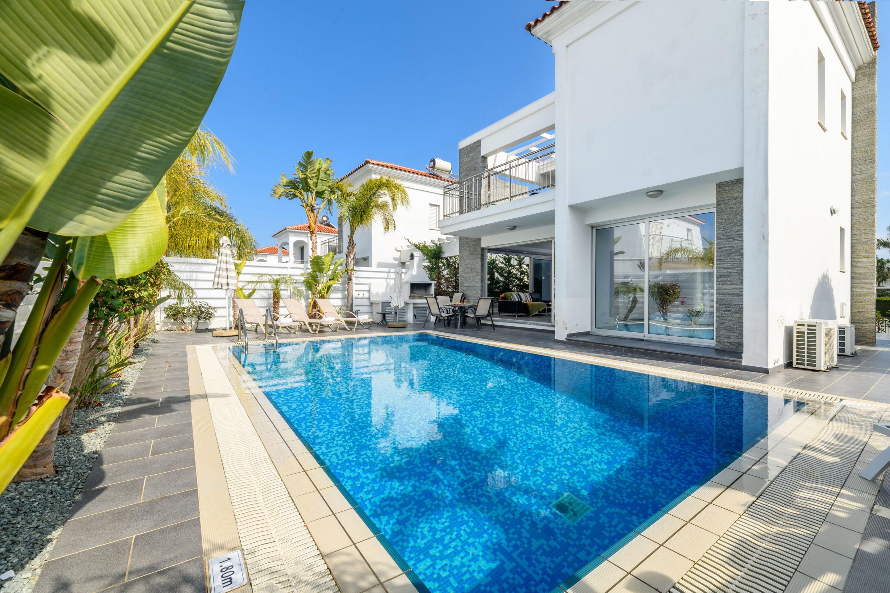 A modern pool area with a clear blue pool, surrounded by white buildings and palm trees.