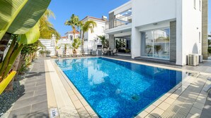 Un área de piscina moderna con una piscina azul cristalina, rodeada de edificios blancos y palmeras.