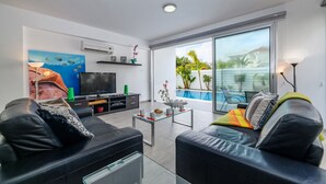 Un salon moderne doté d’un téléviseur à écran plat, de canapés en cuir, d’une table basse en verre et d’une grande fenêtre donnant sur une piscine et un espace verdoyant.