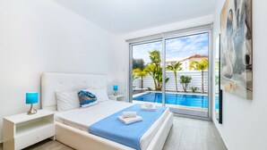 Un dormitorio moderno con una cama grande, una mesita de noche, una lámpara y vista a una piscina a través de un ventanal.