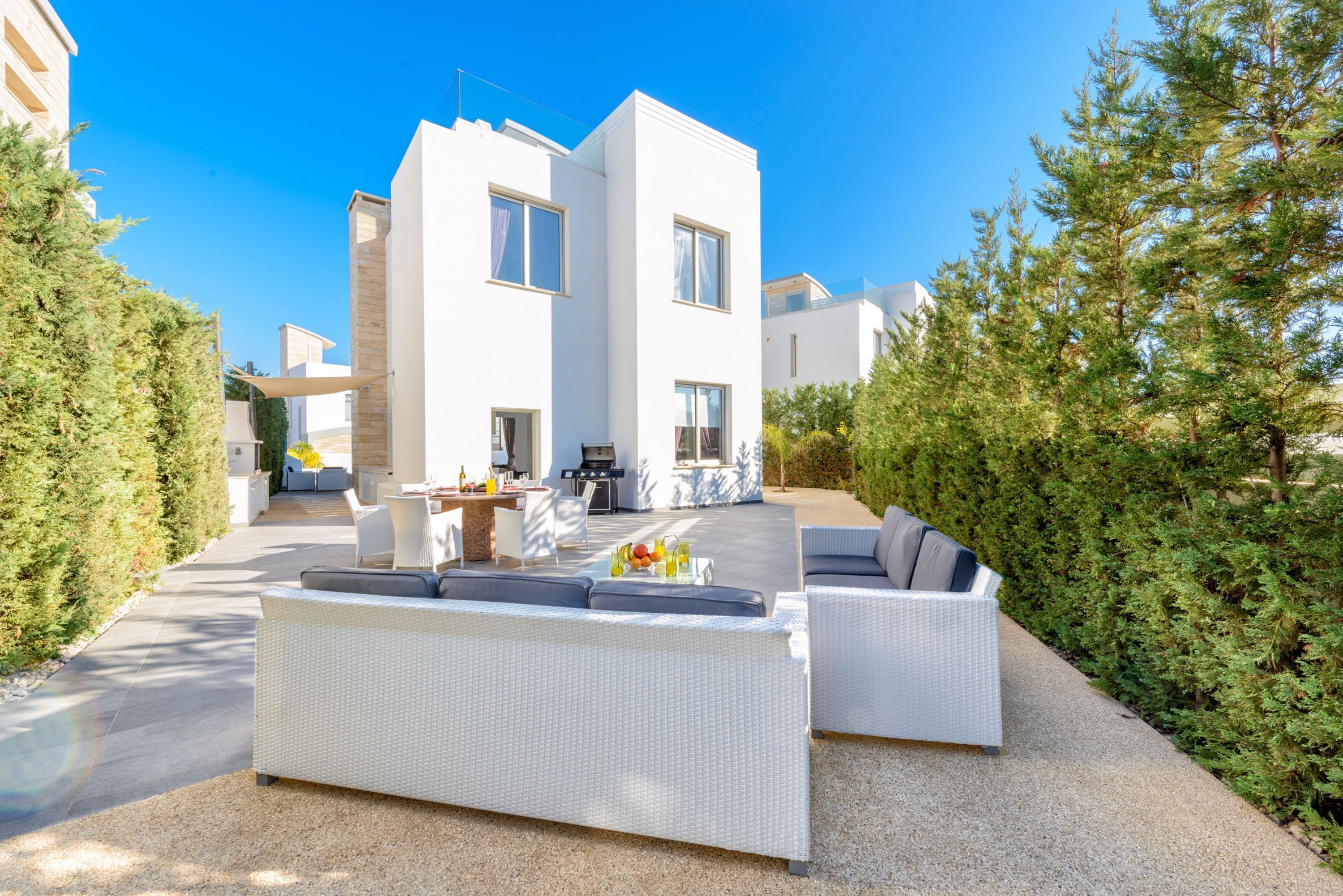 A modern outdoor living area with a white wicker sofa, a dining table with chairs, and a barbecue grill.