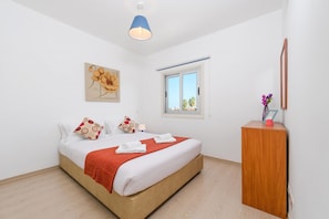 A bedroom with a bed, a wooden nightstand, a window, a ceiling light, and a wall-mounted mirror.