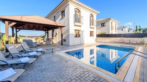 Un espace piscine avec des chaises longues, un coin salon abrité et un bâtiment doté d’un toit en tuiles.