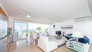 Un salon moderne avec un canapé blanc, une table basse en verre et un téléviseur. Il y a un balcon avec vue sur l’océan.
