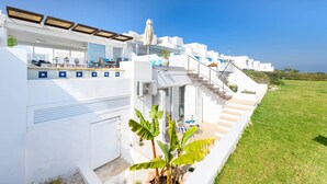 Un bâtiment moderne de couleur blanche, doté d’un espace extérieur aménagé avec des sièges, d’un escalier menant à un balcon et d’une pelouse bien entretenue.