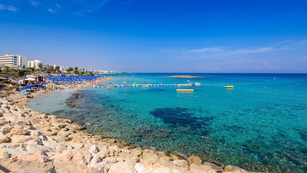 A beach with clear turquoise water, rocky shoreline, and blue umbrellas. - Villa Penelope in Protaras (Paralimni)