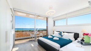 Una habitación de hotel moderna con un gran ventanal corredero que da a un balcón con vistas a la playa. La habitación tiene una cama con edredón azul y blanco, una mesita de noche con lámpara y una pequeña mesa con sillas.