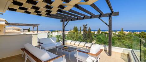Una terraza con mobiliario blanco, una pérgola y vistas al mar.