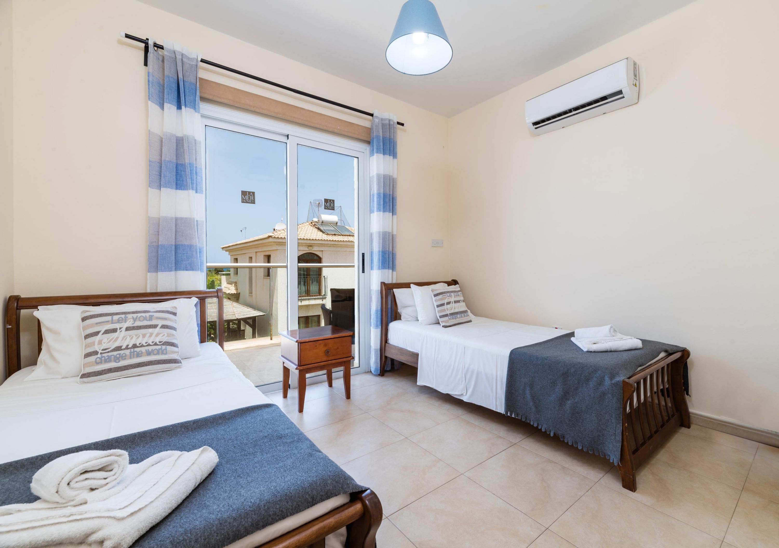 A hotel room with two beds, a small table, a chair, and a view of a building outside.