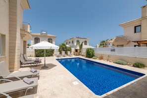 Una piscina con sillas de descanso y sombrillas, rodeada de edificios de arquitectura mediterránea.