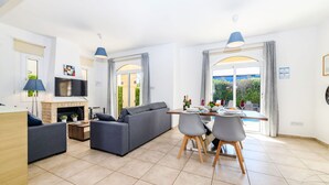 Una sala de estar moderna con sofá, mesa de comedor y sillas, chimenea y ventanas grandes con cortinas.