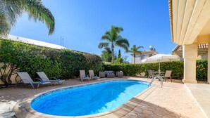 A swimming pool with lounge chairs and umbrellas, surrounded by greenery and a tiled patio. - Villa Oscar in Protaras (Paralimni)