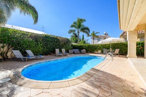 A swimming pool with lounge chairs and umbrellas, surrounded by greenery and a tiled patio.