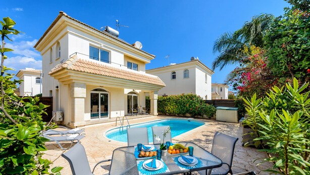 A poolside dining area with a table set for two, surrounded by chairs, and a pool in the background. - Villa Oscar in Protaras (Paralimni)