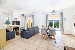 A modern living room with a sofa, dining table, and chairs, a fireplace, and large windows with curtains.