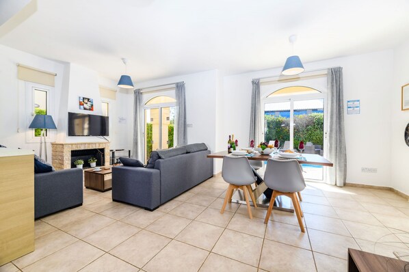 A modern living room with a sofa, dining table, and chairs, a fireplace, and large windows with curtains. - Villa Oscar in Protaras (Paralimni)