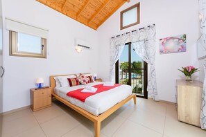 Ein Schlafzimmer mit einem hölzernen Bett, einem Nachttisch mit Lampe, einer Wand-Klimaanlage und Blick ins Freie durch ein großes Fenster.