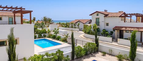 A residential area with two swimming pools surrounded by greenery, white buildings with red-tiled roofs, and a clear view of the sea.