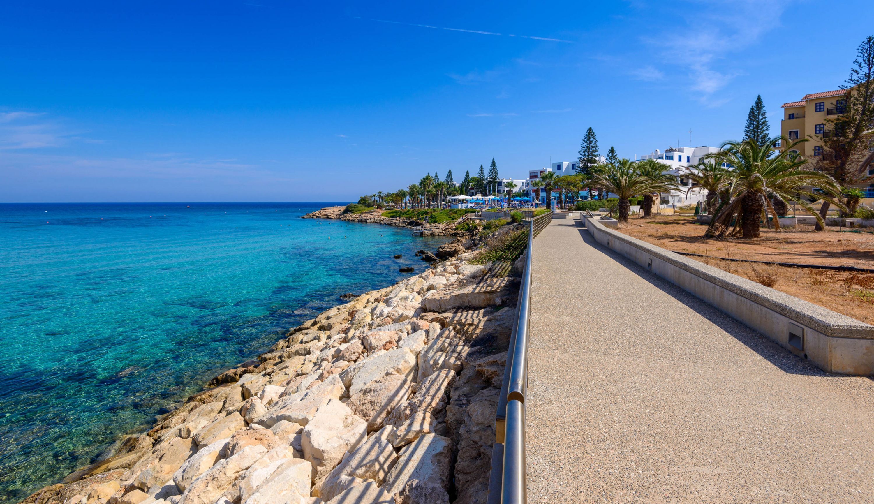 A coastal walkway with a rocky shoreline, clear blue sea, and buildings in the background.