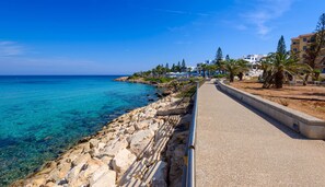 A coastal walkway with a rocky shoreline, clear blue sea, and buildings in the background. - Villa Marilena in Protaras (Paralimni)