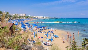A beach with numerous blue umbrellas and sun loungers, a clear blue sea, and buildings in the background. - Villa Marizan in Protaras (Paralimni)