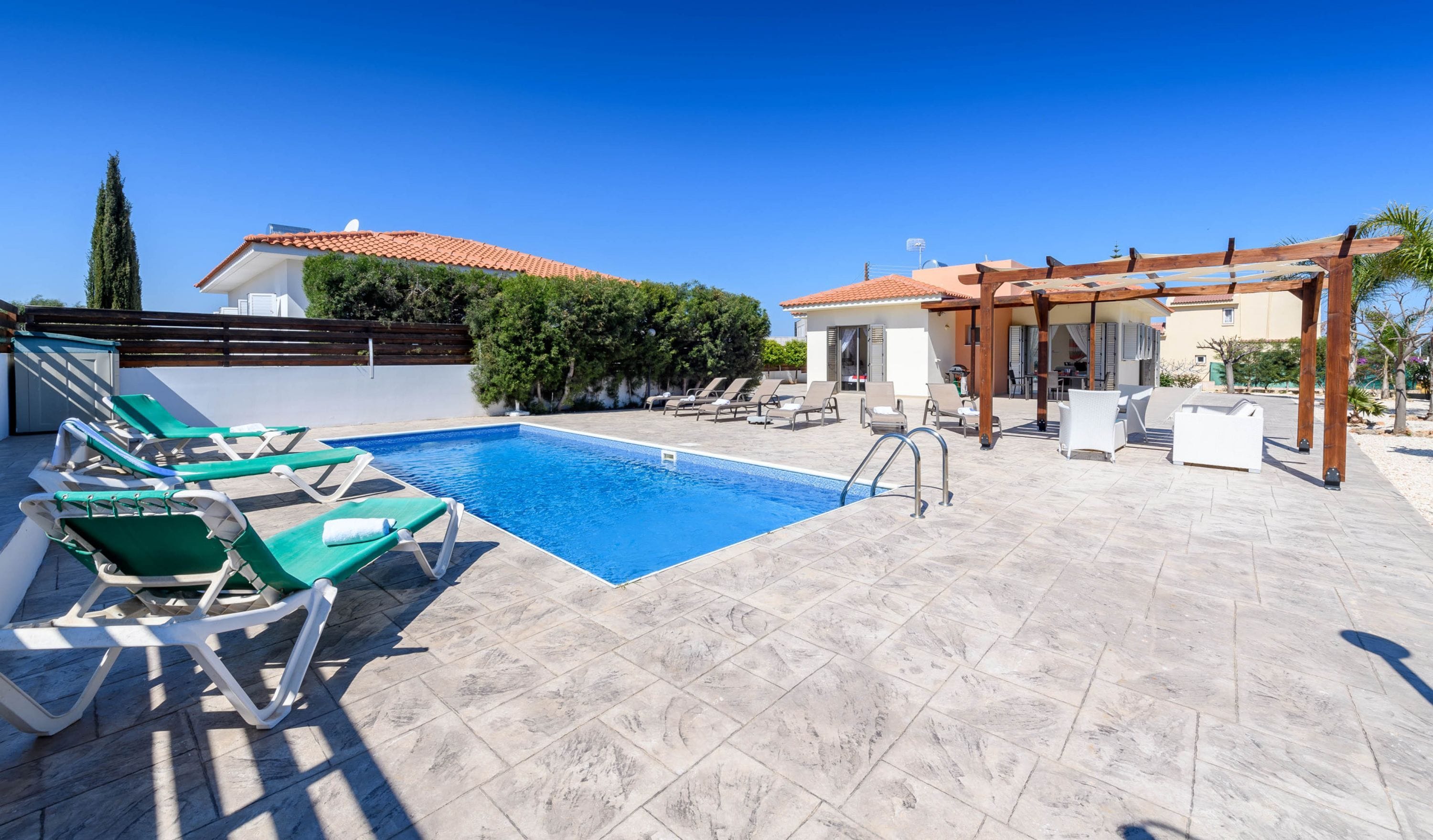 Un espace piscine avec des chaises longues, une pergola et un ciel d’un bleu éclatant.