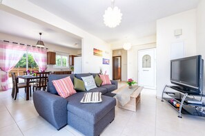 A modern living room with a grey sectional sofa, a dining table with chairs, a TV on a stand, and a kitchen in the background.
