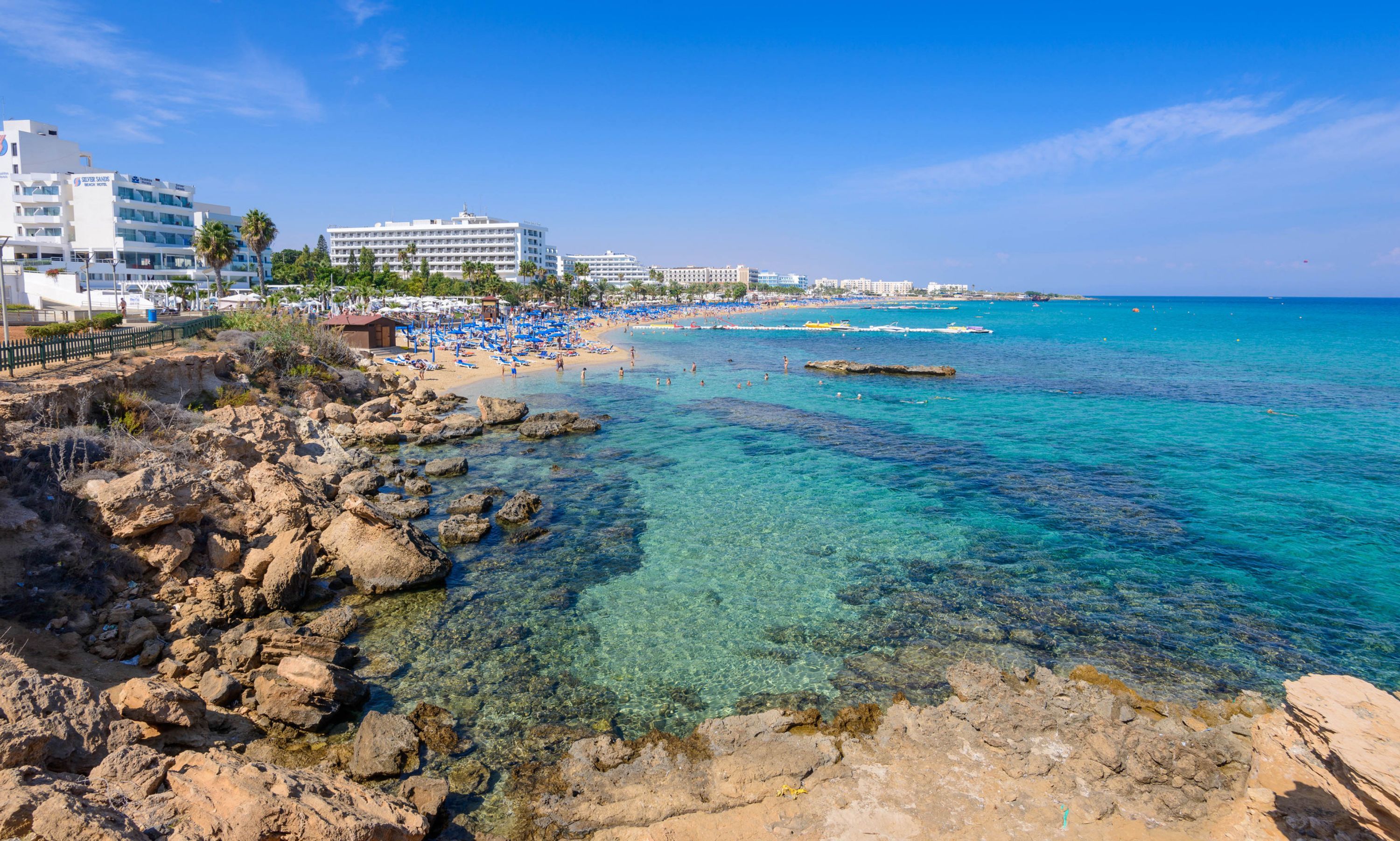 A beach with clear turquoise water, rocky shoreline, and a row of hotel buildings in the background.